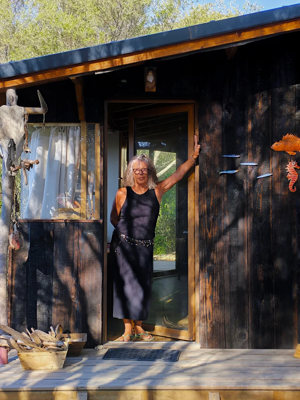 Hélène sur le seuil de la cabane en bois brûlé, sous les arbres.